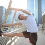 Calm focused African American male athlete with eyes closed in earphones and sportswear leaning on metal fence and doing side bend exercise while listening to music and stretching body on street against blurred urban environment in sunny day