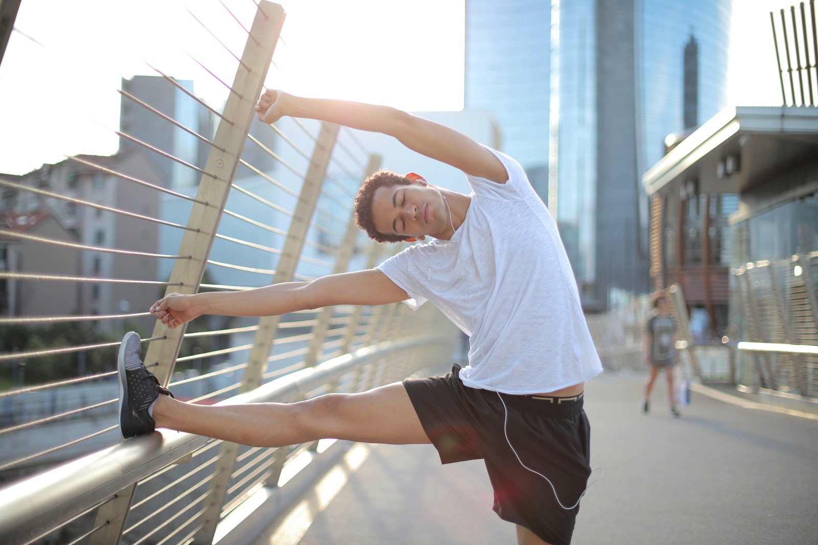 Calm focused African American male athlete with eyes closed in earphones and sportswear leaning on metal fence and doing side bend exercise while listening to music and stretching body on street against blurred urban environment in sunny day